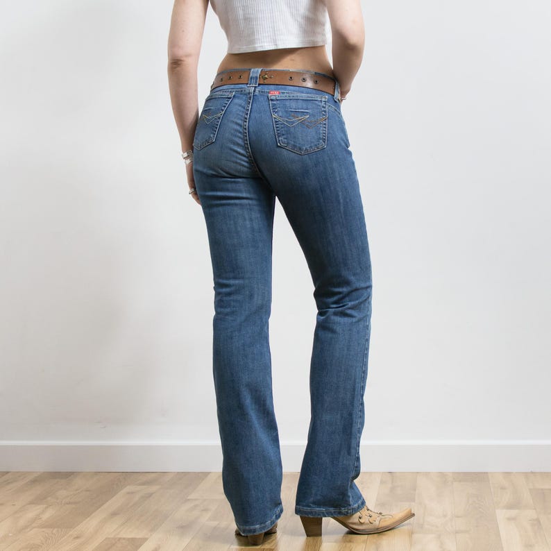 Peut inclure: Jean bleu en denim avec une jambe &eacute;vas&eacute;e, port&eacute; avec une ceinture en cuir marron. Le jean a une conception classique &agrave; cinq poches et est associ&eacute; &agrave; des bottes de cow-boy beiges. Le mod&egrave;le se tient devant un mur blanc et un sol en bois.