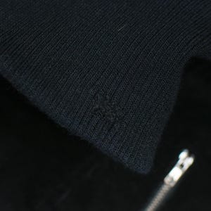 Puede incluir: Primer plano de tejido de punto negro, posiblemente un su&eacute;ter o chaqueta. La tela tiene una textura acanalada y se ve un peque&ntilde;o agujero. Una cremallera plateada es parcialmente visible en la esquina inferior derecha.