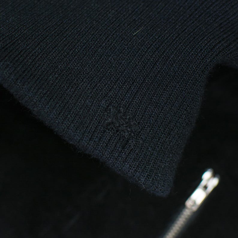 Puede incluir: Primer plano de tejido de punto negro, posiblemente un su&eacute;ter o chaqueta. La tela tiene una textura acanalada y se ve un peque&ntilde;o agujero. Una cremallera plateada es parcialmente visible en la esquina inferior derecha.