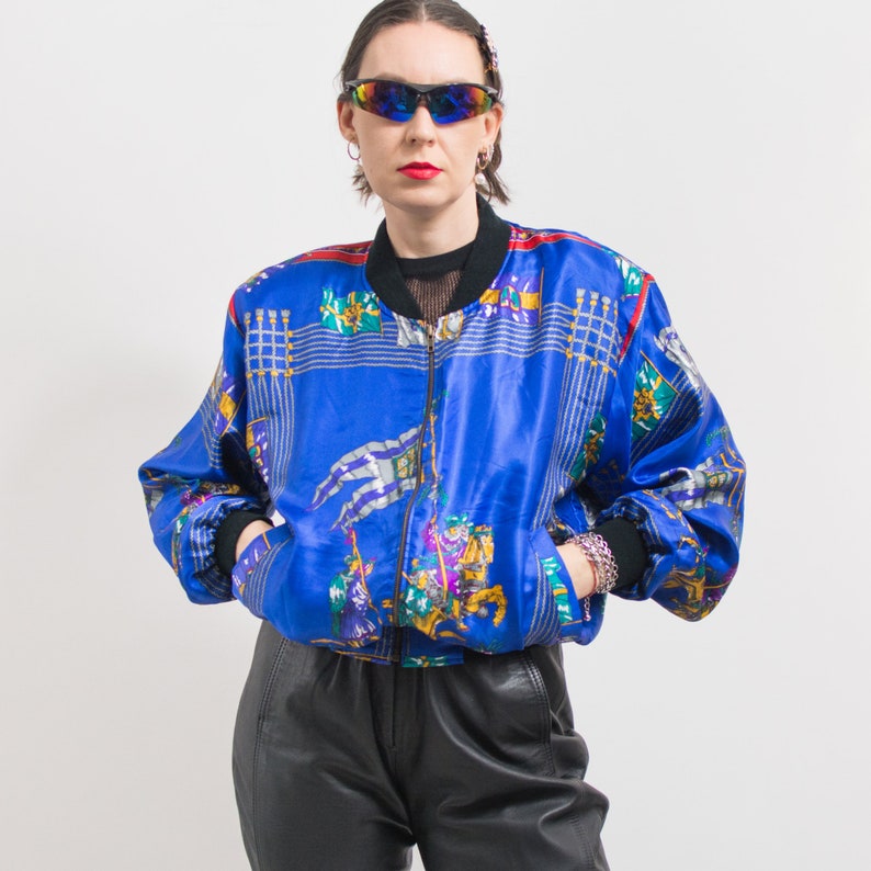 May include: A blue satin bomber jacket with a colorful, abstract print featuring flags and horses. The jacket has a zipper closure and a ribbed collar.