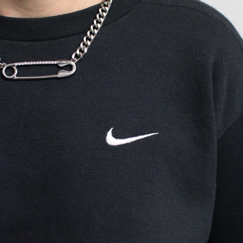 May include: Black crew neck sweatshirt with a white Nike swoosh logo. A silver chain necklace with a large safety pin pendant is worn over the shirt. The safety pin has a row of small, clear stones.