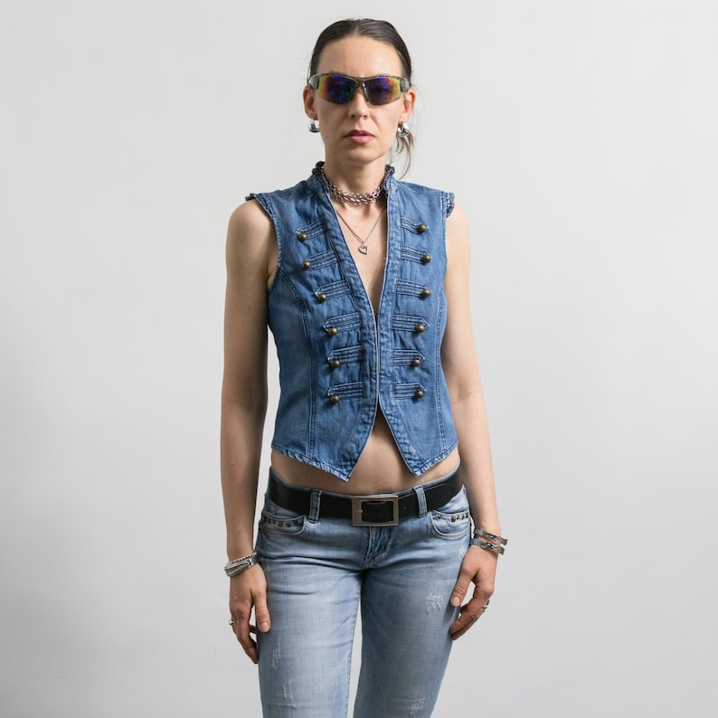 May include: A light blue denim vest featuring a military-inspired design with two vertical rows of brass-toned buttons and decorative cord detailing. The vest is worn with light blue denim jeans, a black leather belt, and reflective sunglasses.