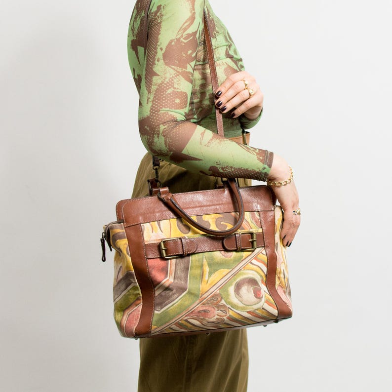 May include: A brown leather and canvas tote bag with a colorful abstract design. The bag features brown leather trim, handles, and straps with metal buckles. The canvas portion displays a mix of yellow, green, red, and brown colors.