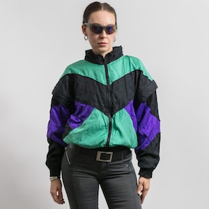 Può includere: Una giacca a vento a blocchi di colore con collo alto e chiusura frontale a zip. Il capo presenta un motivo geometrico in tonalità vivaci di verde, nero e viola, con maniche lunghe e polsini elasticizzati sui polsi.