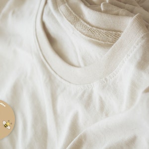 May include: Close-up of several folded, off-white t-shirts. The shirts are made of a soft, textured fabric. A circular sticker in the lower left corner reads "100% Pure Cotton" and "Organic Cotton Shop" with a cotton plant and a bee.