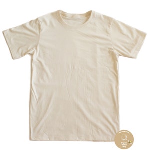 May include: Cream-colored, short-sleeved t-shirt. The shirt has a crew neck and a simple, classic design. A small, circular logo with text is visible in the lower right corner. The shirt appears to be made of a soft, natural fabric.