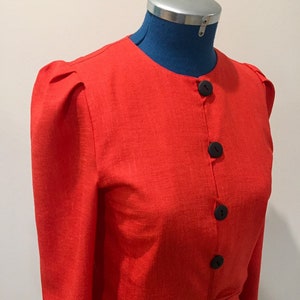 May include: A red linen blazer with long sleeves and a button-up front. The blazer has a round neckline and puffed shoulders.