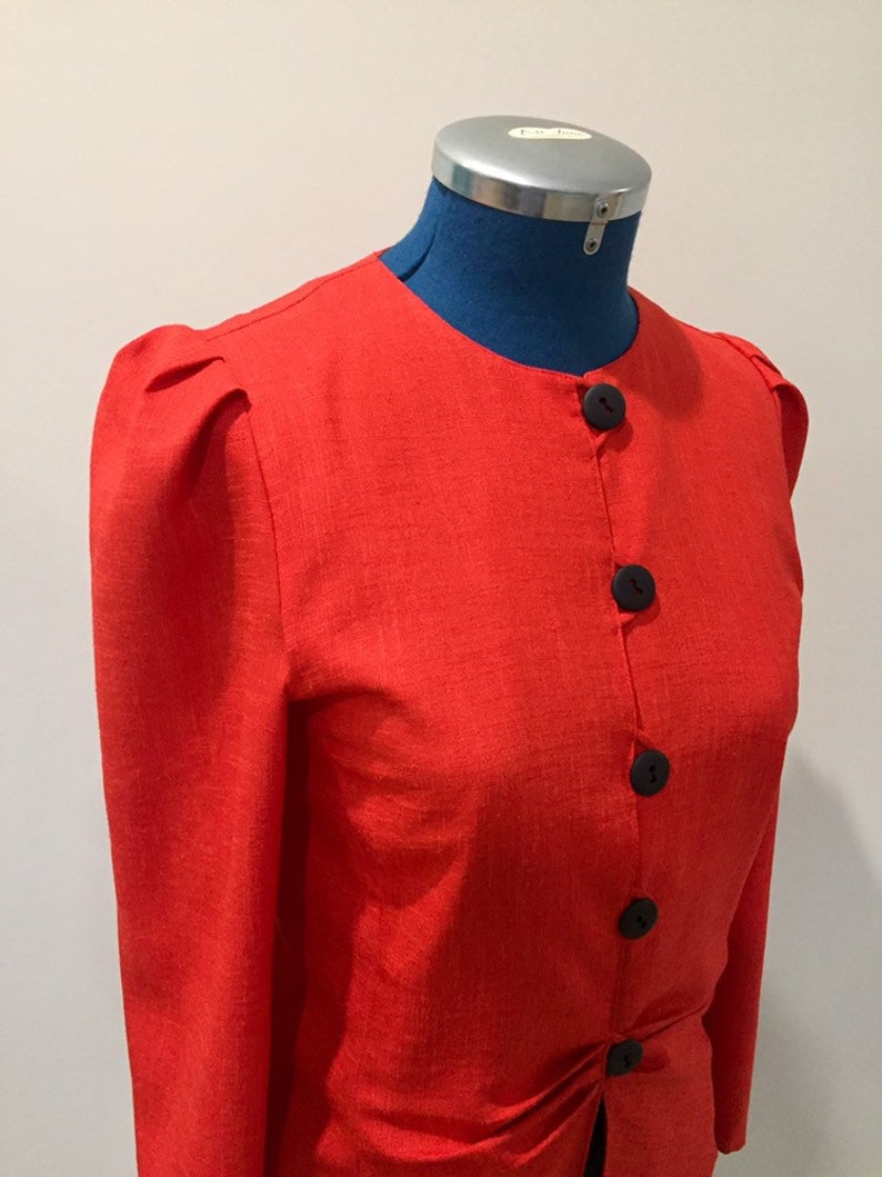May include: A red linen blazer with long sleeves and a button-up front. The blazer has a round neckline and puffed shoulders.