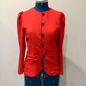 May include: A red button-up jacket with long sleeves and a gathered waist. The jacket has a round neckline and is made of a textured fabric.