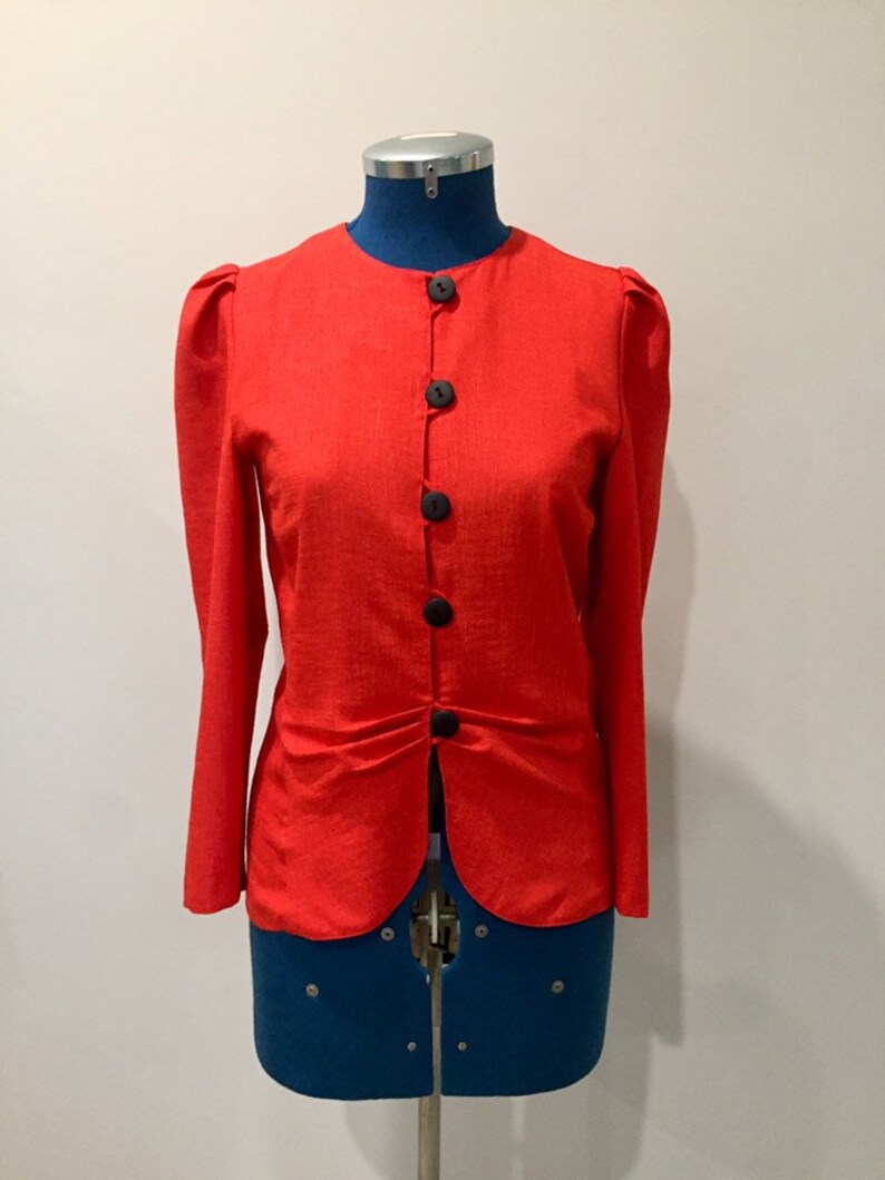 May include: A red button-up jacket with long sleeves and a gathered waist. The jacket has a round neckline and is made of a textured fabric.