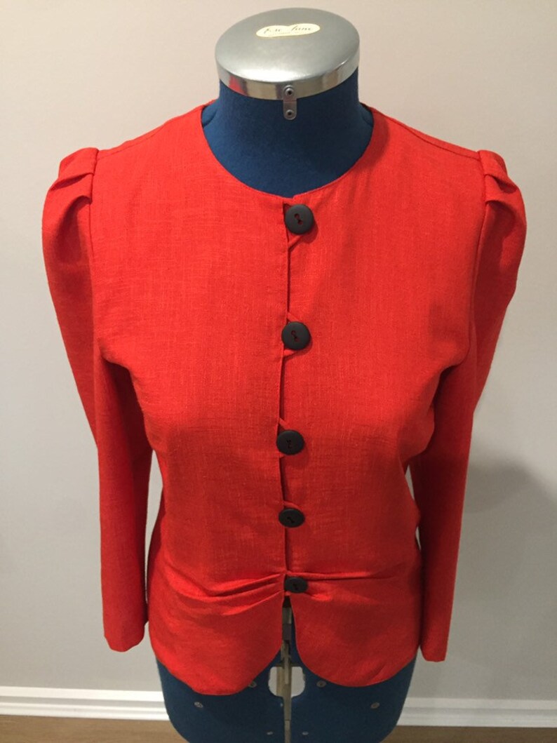 May include: A red linen blazer with a round neckline and five black buttons. The blazer has puffed sleeves and a fitted waist.