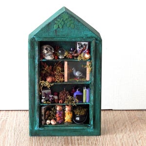 Peut inclure: Une étagère en bois en forme de maison de couleur sarcelle avec trois niveaux contenant des objets miniatures, notamment un crâne, une boule de cristal, une figurine d'oiseau, des petits livres, des flacons en verre et une marmite noire.