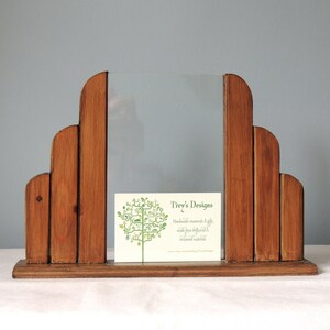 May include: A wooden picture frame with a clear glass insert. The frame is made of multiple pieces of wood that are arranged in a stepped pattern. The frame is sitting on a white surface. A business card with the text "Tivy's Designs" is in the frame.
