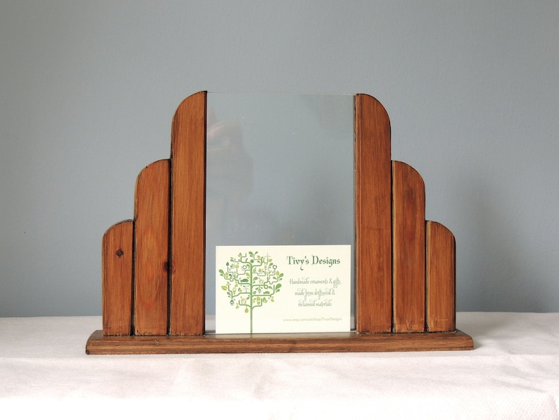 May include: A wooden picture frame with a clear glass insert. The frame is made of multiple pieces of wood that are arranged in a stepped pattern. The frame is sitting on a white surface. A business card with the text "Tivy's Designs" is in the frame.