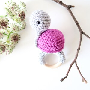 May include: A crocheted turtle rattle toy with a pink shell and gray head and legs. The rattle has a wooden ring in the center.