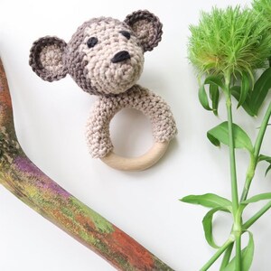 May include: A crocheted monkey rattle with a wooden ring. The monkey is brown and beige with black eyes and a black nose. The rattle is on a white background.