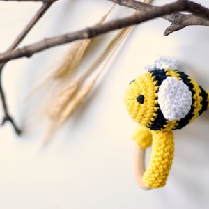 May include: A crocheted bee rattle toy with a yellow and black striped body, white wings, and a wooden ring handle.