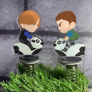 May include: Two acrylic bobblehead figures featuring cartoon characters riding pandas. One character has blonde hair and a blue shirt, while the other has brown hair, a beard, and a green vest. Both figures are mounted on metal springs.