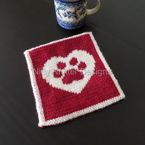 May include: A red knitted coaster with a white paw print inside a heart. The coaster has a white border.