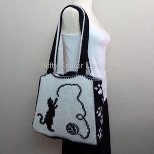 May include: Black and white knitted tote bag with a silhouette of a cat playing with a ball of yarn. The bag has black paw prints on the side.