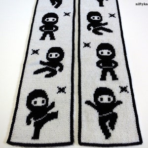 Könnte beinhalten: Ein weißer gestrickter Schal mit schwarzen Ninja-Figuren, die sich über die gesamte Länge des Schals wiederholen. Der Schal hat einen schwarzen Rand.
