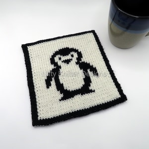 Può includere: Un sottobicchiere quadrato lavorato a maglia con un disegno di pinguino in bianco e nero. Il pinguino è centrato su uno sfondo color crema con un bordo nero. Il testo "Nifty Knitter Designs" è visibile. Una tazza blu e beige è sullo sfondo.