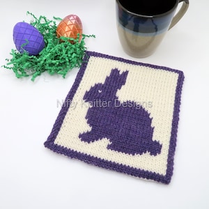 Könnte beinhalten: Ein quadratischer Strickuntersetzer mit einer violetten Hasensilhouette in der Mitte. Der Untersetzer ist weiß mit einem violetten Rand. Der Text "Nifty Knitter Designs" ist in der linken unteren Ecke sichtbar.