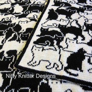 Könnte beinhalten: Schwarze und weiße gestrickte Decke mit einem sich wiederholenden Muster von Katzen. Die Decke ist aus weichem Garn gefertigt und hat eine strukturierte Oberfläche. Der Text "Nifty Knitter Designs" ist in der linken unteren Ecke des Bildes sichtbar.