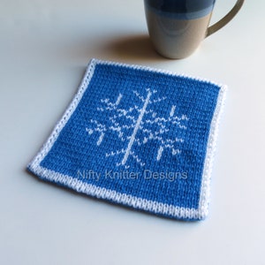 May include: A blue knitted coaster with a white snowflake design. The coaster has a white border and the text "Nifty Knitter Designs" is printed on the bottom.