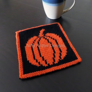 Puede incluir: Un posavasos de calabaza naranja tejido con contorno negro. El posavasos es cuadrado y tiene el texto "Nifty Knitter Designs" en el centro.