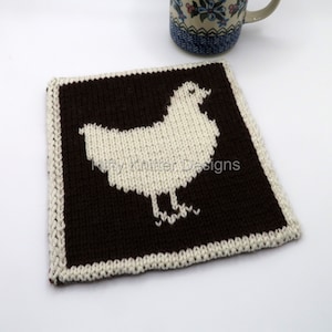 May include: A brown knitted coaster with a white knitted chicken design. The coaster is square and has a white border.