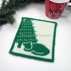 May include: A green and white knitted square featuring a Christmas tree and a cat design. The square has a green border and the text "Nifty Knitter Designs". A red mug with a white snowflake design is in the background.