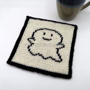 May include: A white knitted square coaster with a black outline and a black and white ghost design in the center. The ghost has a smiling face.