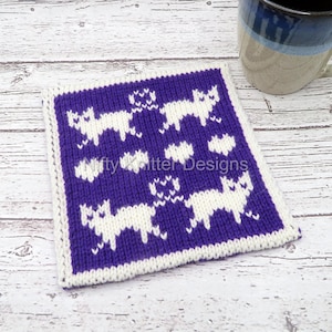 Pode incluir: Uma base de copo quadrada, tricotada, com fundo roxo e padrão de gato branco. A base de copo tem uma borda branca e o texto "Nifty Knitter Designs" é visível. Uma caneca azul e branca está no fundo.