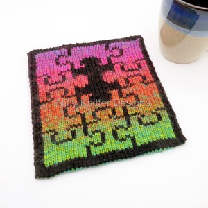 May include: A colorful knitted coaster with a geometric pattern in shades of pink, orange, green, and black. The coaster is made with a black yarn and has a gradient effect. The text "Nifty Knitter Designs" is visible in the center of the coaster.