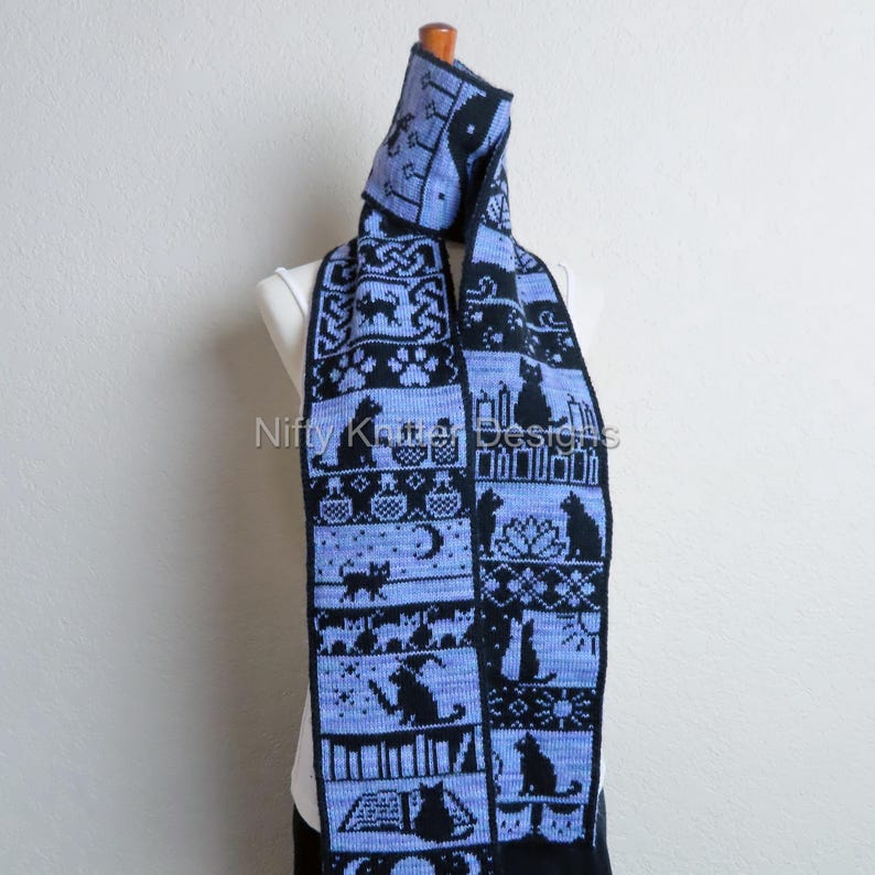 May include: A blue and black knitted scarf with a hood, featuring a repeating pattern of cat silhouettes, candles, and books. The scarf is displayed on a mannequin. The design includes Celtic knots and paw prints. The scarf is approximately 72 inches long.