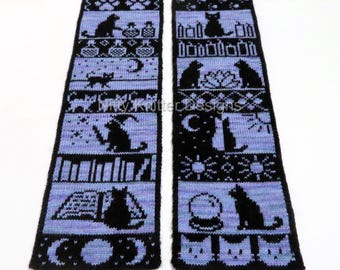 May include: A black and periwinkle knitted scarf featuring repeating patterns of cats, moons, books, and other witchy symbols. The text "Kitty Knitter Designs" is visible on the scarf. The scarf is displayed on a white background.