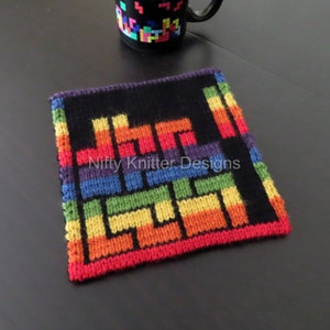 Könnte beinhalten: Ein gestrickter Untersetzer in Regenbogenfarben mit schwarzem Hintergrund und einem Tetris-ähnlichen Muster. Der Untersetzer ist aus verschiedenen Farben gefertigt, darunter Rot, Orange, Gelb, Grün, Blau und Lila. Der Text "Nifty Knitter Designs" ist in der Mitte des Untersetzers sichtbar.