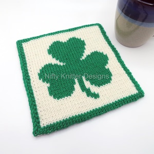 Può includere: Un sottobicchiere di maglia bianca con un design a quadrifoglio verde. Il sottobicchiere è quadrato e ha un bordo verde. Il testo "Nifty Knitter Designs" è visibile al centro del sottobicchiere.
