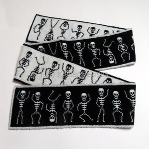 Könnte beinhalten: Ein langer, rechteckiger Strickschal mit einem sich wiederholenden Muster aus tanzenden Skeletten. Das Design wechselt zwischen schwarzen Skeletten auf weißem Grund und weißen Skeletten auf schwarzem Grund, eingefasst mit einem hellgrauen Rand.