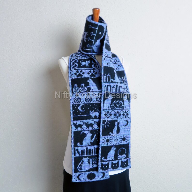 May include: A knitted scarf in black and light blue, featuring a repeating pattern of cats, moons, and other Halloween-themed designs. The scarf is displayed on a mannequin. The text "Nifty Knitter Designs" is visible.