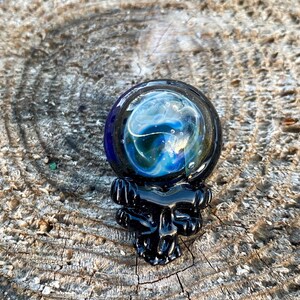 May include: A glass orb with swirling blue and white colors, resting on a black, ornate base. The orb appears to be a decorative item, possibly a paperweight or a small sculpture. The background is a textured, wooden surface.