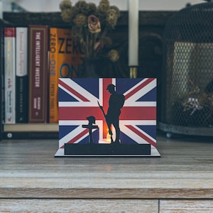 Puede incluir: Una silueta negra de un soldado de pie frente a una cruz con una vela encendida. El soldado está de pie frente a una bandera del Reino Unido. La escena está sobre una superficie de madera.