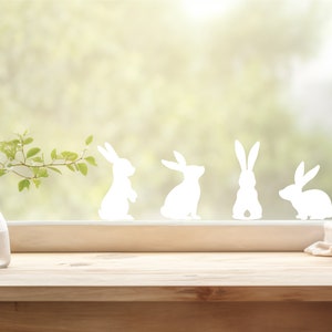 May include: A window with a white wooden frame and a view of green trees and bushes. There are five white bunny silhouettes on the window. A wooden countertop with two glass vases and a white plate with apricots on it.