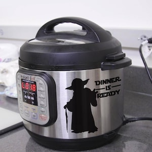 Può includere: Una pentola a pressione Instant Pot in acciaio inossidabile con un coperchio nero e un adesivo con la silhouette di Yoda. L'adesivo dice "Dinner is Ready".