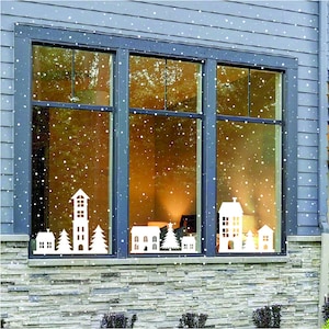 May include: A window with a dark frame is decorated with white die-cut village silhouettes and falling snow. The silhouettes include houses, a tall tower, and evergreen trees. The window is set in a blue-gray house exterior with a stone base.