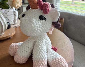 Könnte beinhalten: Ein handgefertigtes Häkel-Einhorn-Stofftier mit weißem Körper, beigem Horn und einer Mähne aus rosa und dunkelbraunem Garn. Das Wesen hat große schwarze Sicherheitsaugen aus Kunststoff und hellrosa Akzente an den Spitzen der vier Beine.