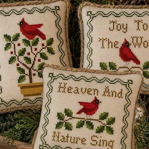 May include: Three square fabric ornaments with cross-stitch designs featuring red cardinals, green holly leaves, and tan borders. One shows a cardinal in a pot, another says Joy To The World, and the third says Heaven And Nature Sing in brown text.