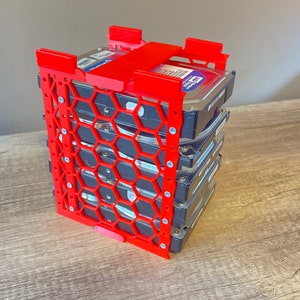 以下が含まれることがあります： ハニカムデザインの赤い3Dプリントハードドライブラックに、ハードドライブのスタックが収納されています。ハードドライブは黒で、青いラベルには「Western Digital」と書いてあります。
