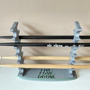 K&ouml;nnte beinhalten: Ein grauer 3D-gedruckter Schlagzeugst&ouml;ckehalter mit drei Paaren Schlagzeugst&ouml;cken. Der Halter hat die Worte "Live Love Drum" auf der Basis aufgedruckt. Der oberste Schlagstock ist ein schwarzes Vic Firth Signature-Modell.